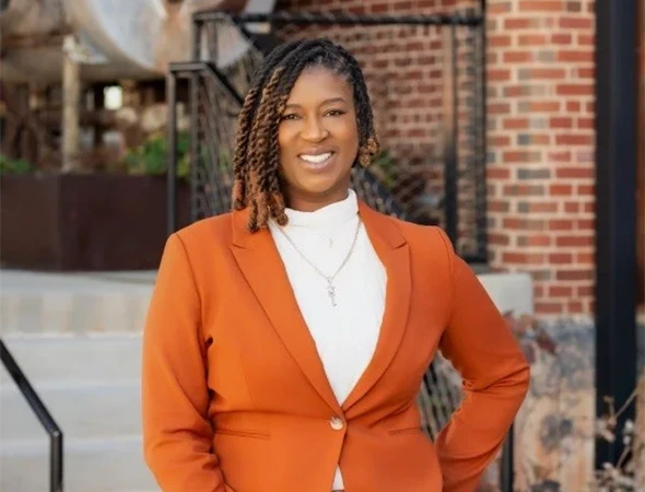 Portrait of the CEO of Another Chance Bail Bonds, a woman with dreadlocks wearing an orange blazer and a white turtleneck, standing outdoors in front of a brick building.