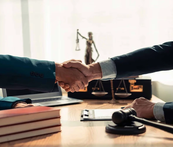 Two people in business suits shaking hands across a desk, with a bronze Lady Justice statue visible in the background.